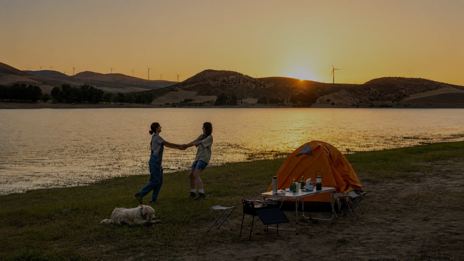 Two people holding hands near a campsite by a lake at sunset, with an orange tent, folding table, chairs, and a dog on the grassy shore. Hills and a glowing sun are visible in the background.