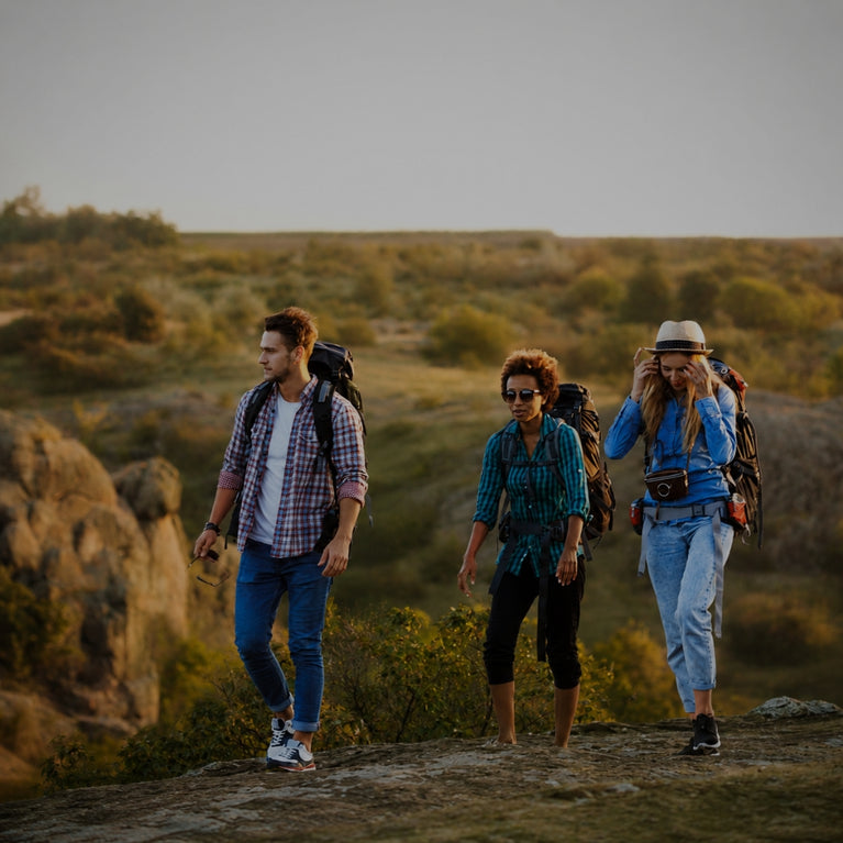 Three hikers with backpacks walking along a rocky trail in a sunlit outdoor landscape with shrubs and rock formations.