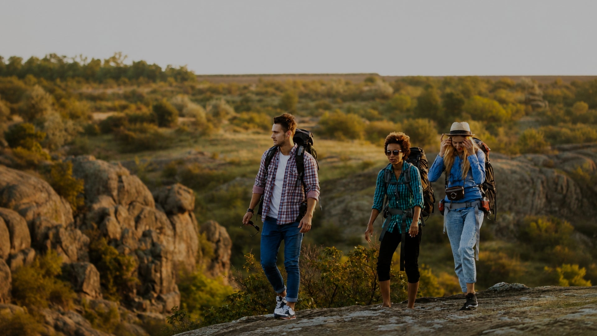 Three hikers with backpacks walking along a rocky trail in a sunlit outdoor landscape with shrubs and rock formations.